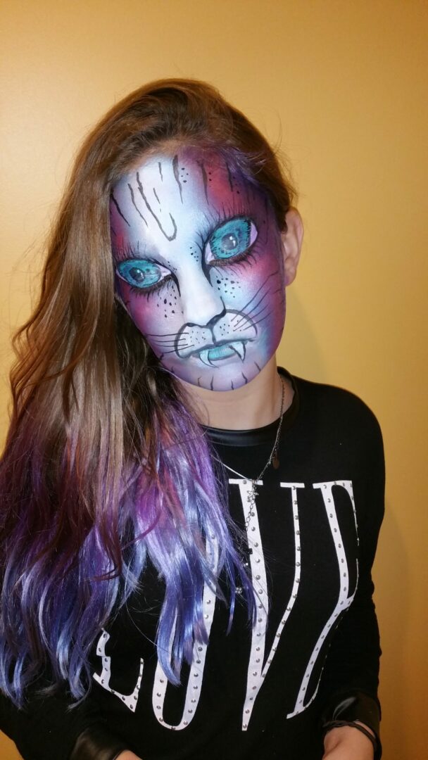 A girl with purple hair and blue eyes has a cat face paint on.